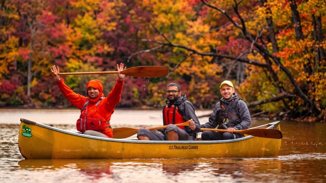 Three people in a canoe holding their paddles above their head against a fall colour backdrop